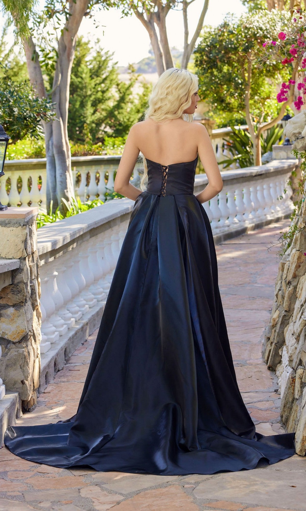 A blonde woman stands outdoors on a stone path, wearing the Juliet JT2733H—a strapless black organza a-line prom dress with a lace-up back and sweeping train.